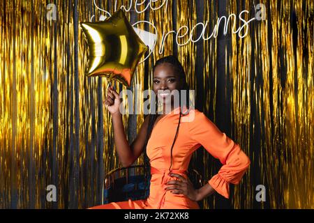 Joyeux jeune femme afro-américaine souriante avec des dreadlocks dans la robe orange tenir le ballon étoile dorée de fête. Doux rêve Banque D'Images