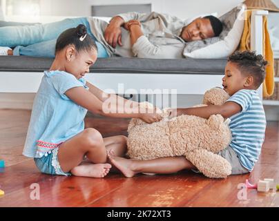 Pendant que vous dormiez. deux frères et sœurs se battent sur un teddy sur le sol tandis que leur père dort sur le canapé à la maison. Banque D'Images