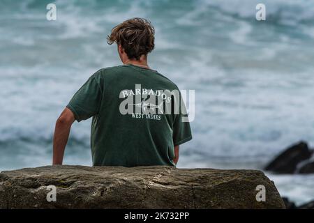 Un jeune homme assis seul sur un rack et portant un T-shirt vert avec le mot Barbade et un logo sur le dos. Banque D'Images