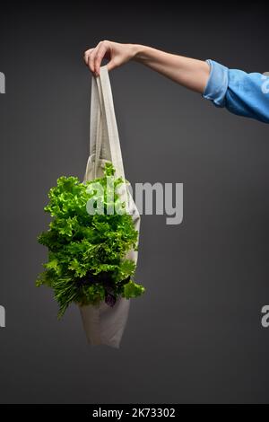 Main de femme dans une chemise en denim bleu contenant un sac à provisions réutilisable avec bouquet de feuilles de salade vertes, basilic, romarin et baume de citron sur le backgro gris foncé Banque D'Images
