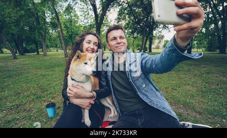 Le couple heureux prend le selfie dans le parc et regarde le smartphone, tandis que la jeune femme porte et caresse le chiot shiba inu race mignon. Banque D'Images