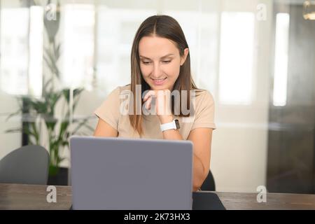 Portrait d'une jeune femme assise au bureau avec un ordinateur portable, saisie d'un e-mail, messagerie d'un ami, chat, flirt, rencontres en ligne avec un sourire agréable Banque D'Images