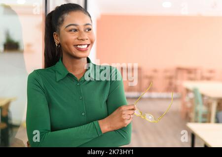 Jeune femme entrepreneure ou femme d'affaires confiante et prospère, vêtue de vêtements décontractés, les bras repliés au bureau et regardant loin tout en tenant des lunettes Banque D'Images
