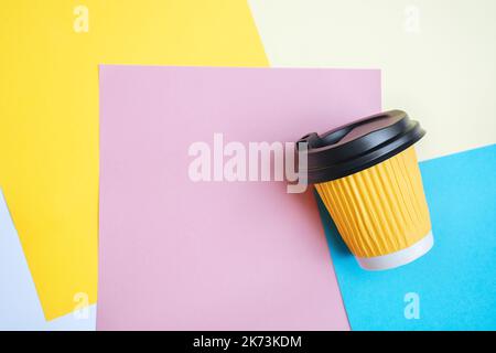 La mise au point sélective douce sur une tasse de café à emporter se trouve dans un désordre créatif sur du papier coloré sur la table de paramétrage. Fond bleu, orange et rose. Copier l'espace pour le texte Banque D'Images