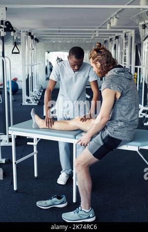 Jeune thérapeute africain massant la jambe du patient avant l'entraînement sportif en salle de gym Banque D'Images