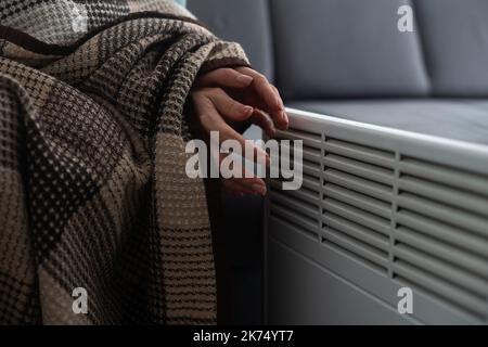 Une fille chauffe les mains gelées au-dessus du radiateur chaud, en gros plan. Femme portant un chandail en laine se réchauffant tout en étant assise près d'un radiateur de chauffage. Femme Banque D'Images