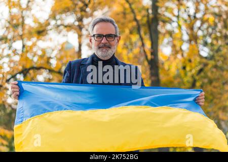 Soutien à l'Ukraine. Confiant, souriant, d'âge moyen, barbu, homme aux cheveux gris tenant le drapeau ukrainien bleu et jaune devant lui de deux mains. Arbres flous en arrière-plan. Photo de haute qualité Banque D'Images