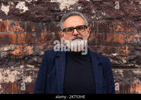 Moderne, confiant, gris-cheveux, homme d'âge moyen dans un blazer et un col roulé, ayant des lunettes sur, debout devant un mur de briques, regardant la caméra. Photo de haute qualité Banque D'Images