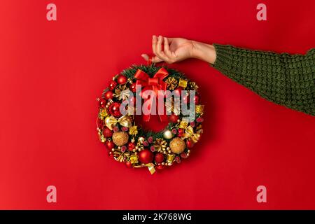 Décoration de fête minimaliste, couronne décorative de Noël entre les mains des femmes sur fond rouge. Arrière-plan lumineux et festif avec espace de copie Banque D'Images