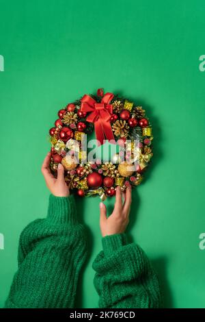 Couronne de sapin de Noël avec décoration festive entre les mains des femmes sur fond vert. Le minimalisme, le concept de préparation pour Noël et le nouvel an Banque D'Images