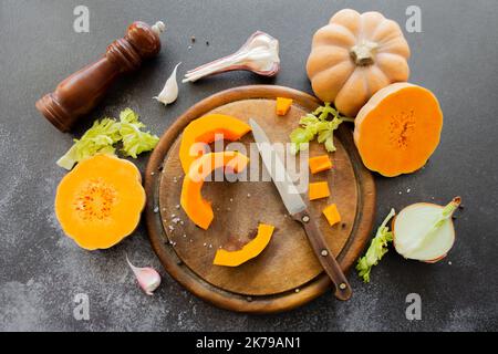 Potiron frais et brillant sur une planche à découper en bois avec un couteau. Céleri et ail, oignons et grains de poivre épars et sel avec moulin à épices. Banque D'Images