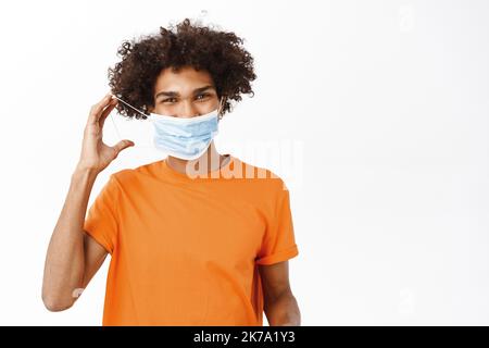 Image d'un beau jeune homme souriant, montrant comment porter un masque médical pendant une pandémie de covid-19, debout dans une chemise orange sur fond blanc Banque D'Images