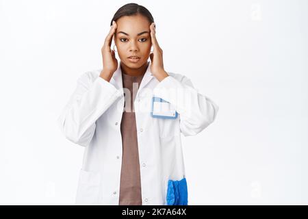 Thérapeute, femme médecin tenant la tête et regardant concerné, ayant des maux de tête, sentir la fatigue, debout sur fond blanc Banque D'Images