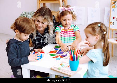 Petits enfants et éducateur qui étudient, plient des cubes et des figures géométriques en papier sur le bureau assis au sol dans la salle de jeux. Leçon intéressante pour Banque D'Images