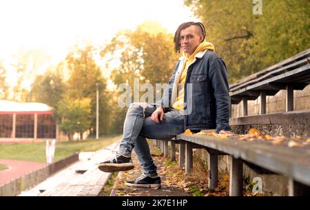 Un jeune homme positif avec des dreadlocks regarde la caméra sur un vieux banc de rue de bleacher en automne doré Banque D'Images