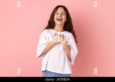 Portrait d'une petite fille folle et optimiste portant un T-shirt blanc écoutant une blague drôle, riant à voix haute, tenant le ventre, exprimant des émotions positives. Studio d'intérieur isolé sur fond rose. Banque D'Images