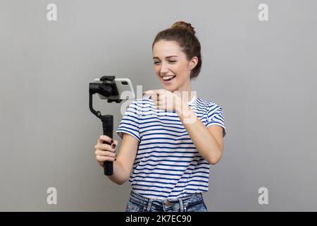 Portrait d'une femme attrayante portant un T-shirt rayé pointant vers l'appareil photo de téléphone portable avec le sourire, streaming, parlant avec des suiveurs, utilise Steadicam. Prise de vue en studio isolée sur fond gris. Banque D'Images