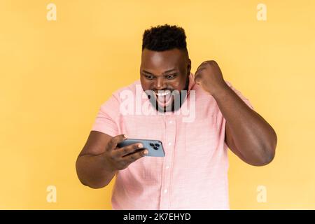 Portrait d'un homme portant une chemise rose debout, utilisant un smartphone et jouant à un jeu mobile avec le visage positif excité, heureux de gagner, poing serré. Studio d'intérieur isolé sur fond jaune. Banque D'Images