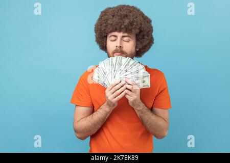 Portrait d'un homme avec une coiffure afro portant un T-shirt orange qui sent des billets de banque en dollars avec plaisir, profitant de gros profits et de la vie riche. Studio d'intérieur isolé sur fond bleu. Banque D'Images
