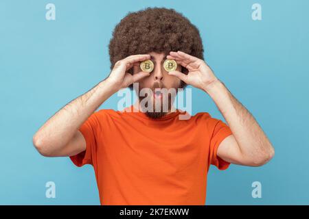 Portrait d'un homme étonné avec une coiffure afro portant un T-shirt orange couvrant les yeux avec du bitcoin doré, regardant l'appareil photo à travers la pièce de btc. Studio d'intérieur isolé sur fond bleu. Banque D'Images