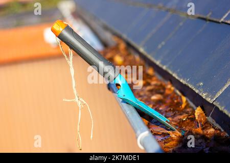 Un portrait d'une gouttière de toit pleine de feuilles d'automne colorées tombées un jour ensoleillé avec une petite pelle de jardin bleue dans elle de quelqu'un qui nettoie le Banque D'Images