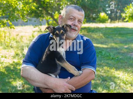 Joli portrait positif de l'homme ukrainien senior assis dans le jardin d'été avec un chiot doux sur les mains sorcière est dans un endroit de sauvegarde. Banque D'Images