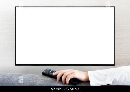 Télécommande de téléviseur dans la main d'un homme devant le téléviseur grand écran avec écran blanc sur fond de mur blanc. Guy bascule les chaînes de la télécommande depuis le Banque D'Images