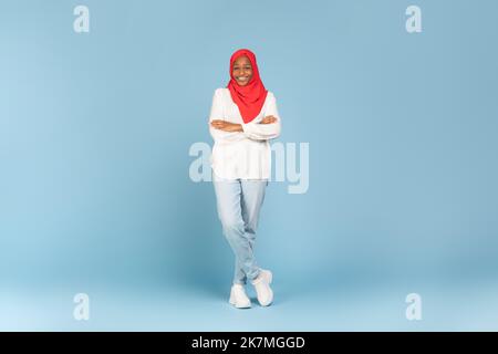Prise de vue en longueur d'une femme afro-américaine heureuse dans le hijab posant avec les bras pliés sur fond bleu, espace libre Banque D'Images