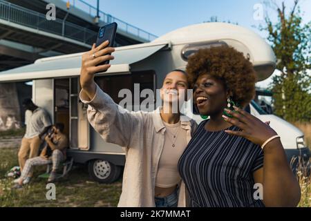 Deux belles personnes en premier plan - queer personne caucasienne prenant un selfie avec le téléphone portable noir avec leur ami noir sympathique en robe dépouillée. Cabine de camping et amis masculins avec guitare en arrière-plan. Photo de haute qualité Banque D'Images