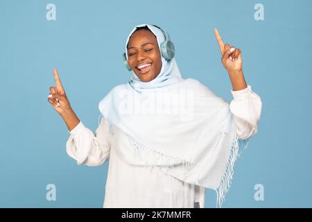 Une femme musulmane afro-américaine surjoyée dans le hijab s'amusant, dansant et chantant avec des mains levées, fond bleu Banque D'Images