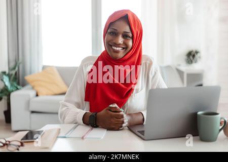 Portrait d'une femme indépendante musulmane noire en hijab assise à table avec un ordinateur portable et souriant à la caméra, travaillant à domicile Banque D'Images