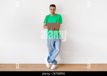 Programmes d'éducation en ligne. Un homme indépendant arabe heureux avec un ordinateur portable dans les mains, debout sur un mur léger, plein plan Banque D'Images