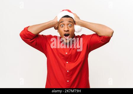 Un gars afro-américain choqué en chapeau de père noël et un maillot rouge tenant la tête avec les deux mains isolées sur fond blanc, un gars étonné regarde la caméra avec les yeux et la bouche ouverts, une promotion incroyable Banque D'Images