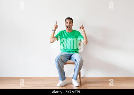 Homme arabe surjoyé assis sur une chaise et pointant deux doigts vers le haut, offrant de l'espace pour votre publicité, mur léger Banque D'Images