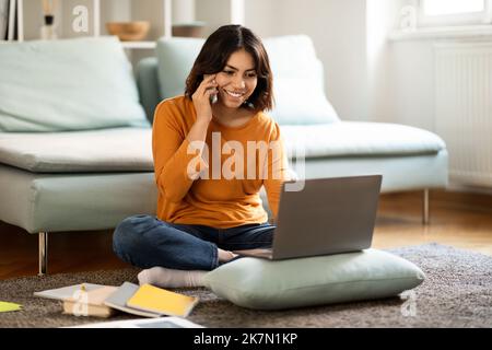 Femme arabe souriante parlant au téléphone portable et travaillant avec un ordinateur portable à la maison Banque D'Images