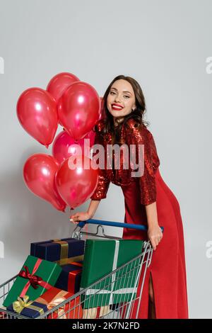 Femme heureuse et élégante debout près du panier avec des ballons et des cadeaux sur fond gris, image de stock Banque D'Images