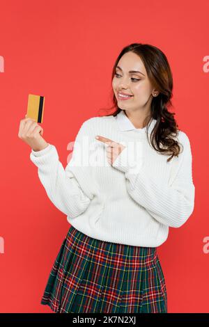 Femme souriante en jupe à carreaux et chandail pointant à la carte de crédit isolée sur rouge, image de stock Banque D'Images