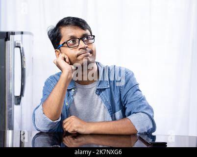Portrait d'un jeune homme indien d'âge moyen portant des lunettes assis à la table et pensant vers le haut sur fond blanc à la maison Banque D'Images