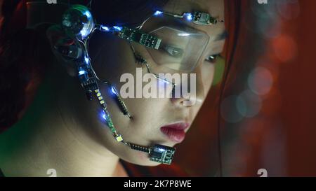 Une fille asiatique concentrée dans la cyberpunk attyre travaille sur l'ordinateur. Porter des lunettes à un œil avec des LED blanches et un microphone. Arrière-plan des lumières néon. science fiction, concept de cybernétique. Banque D'Images