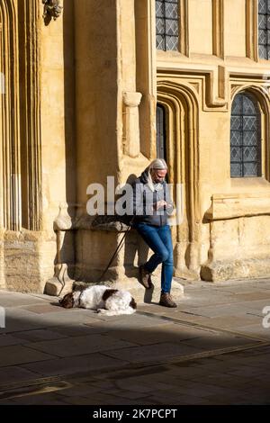 Une femme mûre utilisant son téléphone penchée contre l'entrée de St. Église Jean-Baptiste à Cirencester (Oct22) Banque D'Images