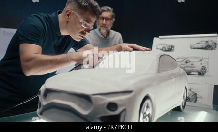 L'ingénieur automobile travaille sur la sculpture d'un prototype de voiture à l'aide d'un outil rotatif tout en discutant avec son collègue principal. Entreprise de conception de voitures de haute technologie avec des technologies innovantes. Banque D'Images