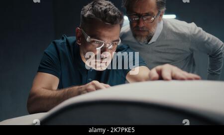 Un ingénieur concepteur équipé de lunettes de sécurité travaille sur la sculpture d'un prototype de voiture à l'aide d'un outil de type stylo. Apporter des corrections à la conception et la perfectionner tout en discutant avec un collègue. Banque D'Images