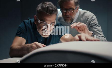 Un ingénieur concepteur équipé de lunettes de sécurité travaille sur la sculpture d'un prototype de voiture à l'aide d'un outil de type stylo. Apporter des corrections à la conception et la perfectionner tout en discutant avec un collègue. Banque D'Images