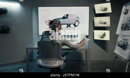 Vue arrière d'un graphiste automobile dessine un croquis du modèle prototype d'une voiture à l'aide d'un stylet dans un laboratoire innovant de haute technologie. Industrie automobile. Studio moderne avec LED. Banque D'Images