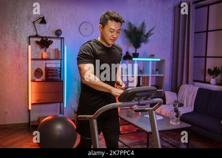 Portrait de l'homme asiatique actif dans les vêtements de sport en utilisant le tapis roulant à la maison la nuit. Coronavirus Covid 19 distance sociale. Entraînement à domicile, sports, adulte actif, séjour à la maison, concept de loisirs à distance. Banque D'Images