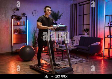 Portrait d'un jeune homme d'affaires actif et dynamique avec casque et papiers faisant du sport fitness à la maison courir sur un tapis roulant à l'intérieur la nuit. Tonifiez votre corps. Forme parfaite. Amélioration de l'endurance Banque D'Images