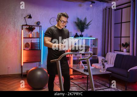 Portrait d'un jeune homme d'affaires asiatique actif dans des vêtements de sport gris et de formation de casque à la maison travaillant avec des papiers, faisant l'exercice cardio sur le tapis roulant à l'heure du soir. Concept de sport, affaires. Banque D'Images