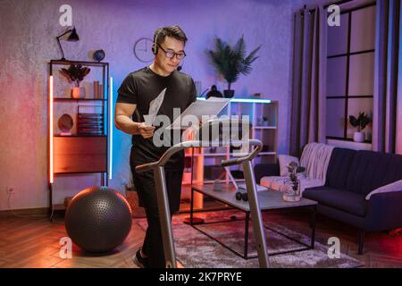 Portrait d'un jeune homme d'affaires asiatique actif dans des vêtements de sport gris et de formation de casque à la maison travaillant avec des papiers, faisant l'exercice cardio sur le tapis roulant à l'heure du soir. Concept de sport, affaires. Banque D'Images