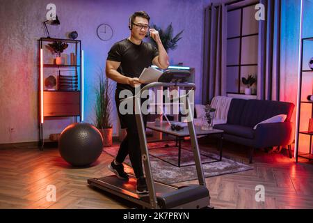 Portrait d'un jeune homme d'affaires actif et dynamique avec casque et papiers faisant du sport fitness à la maison courir sur un tapis roulant à l'intérieur la nuit. Tonifiez votre corps. Forme parfaite. Amélioration de l'endurance Banque D'Images