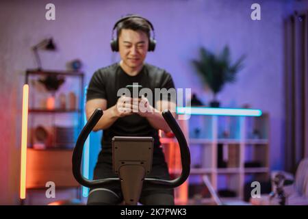 Jeune homme asiatique avec casque faisant des exercices sur vélo stationnaire à la maison ou au centre de remise en forme. Jeune homme sportif en salle de sport écoutez de la musique depuis votre smartphone. Homme faisant des exercices cardio. Banque D'Images